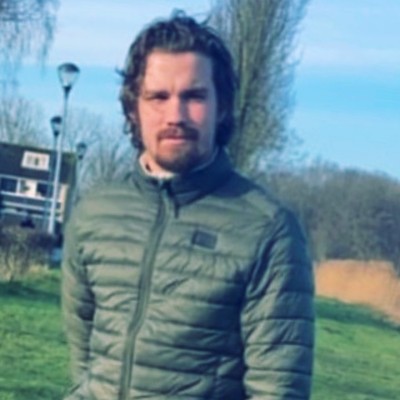 Een man die buiten staat, gekleed in een groene jas, met op de achtergrond gras en bomen.